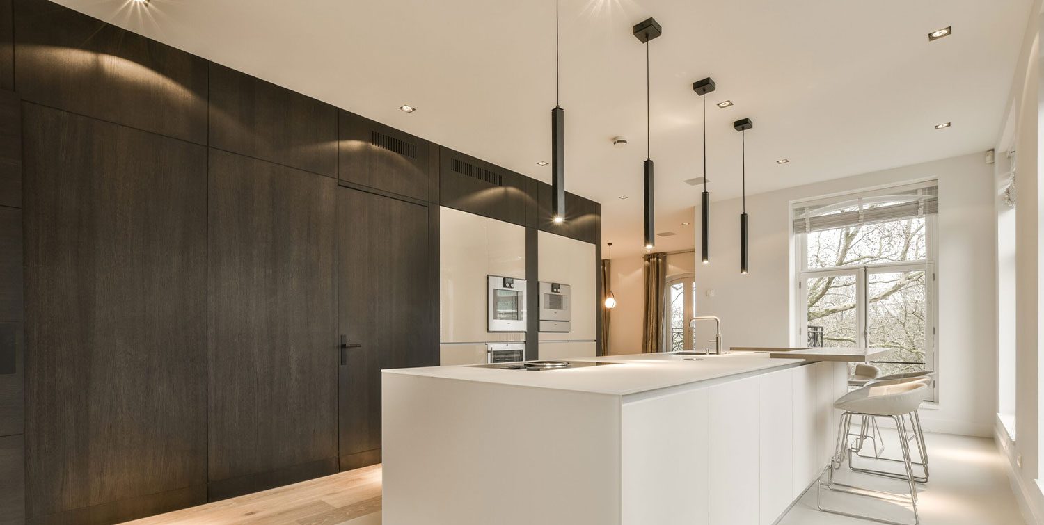 A modern kitchen features a large white island with barstools, built-in ovens, and sleek dark cabinetry. Pendant lights hang from the ceiling providing illumination over the countertops.